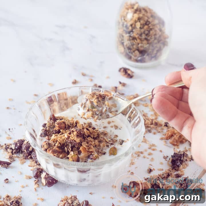 A square photo with a glass bowl with yogurt and chocolate muesli. A golden spoon takes a bite on a marble white background. In the background a glass jar with more chocolate nut granola.