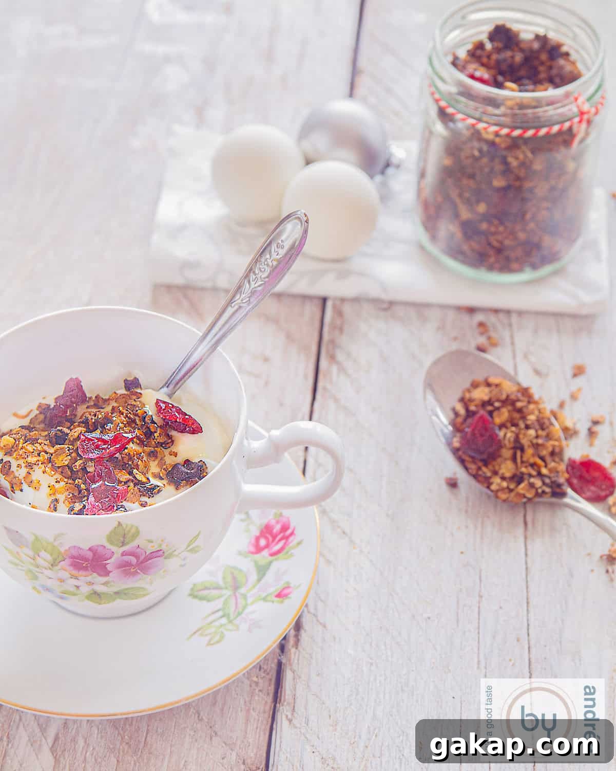 A cup on a plate filled with Christmas granola. In the background a jar with extra granola and a spoon filled with granola