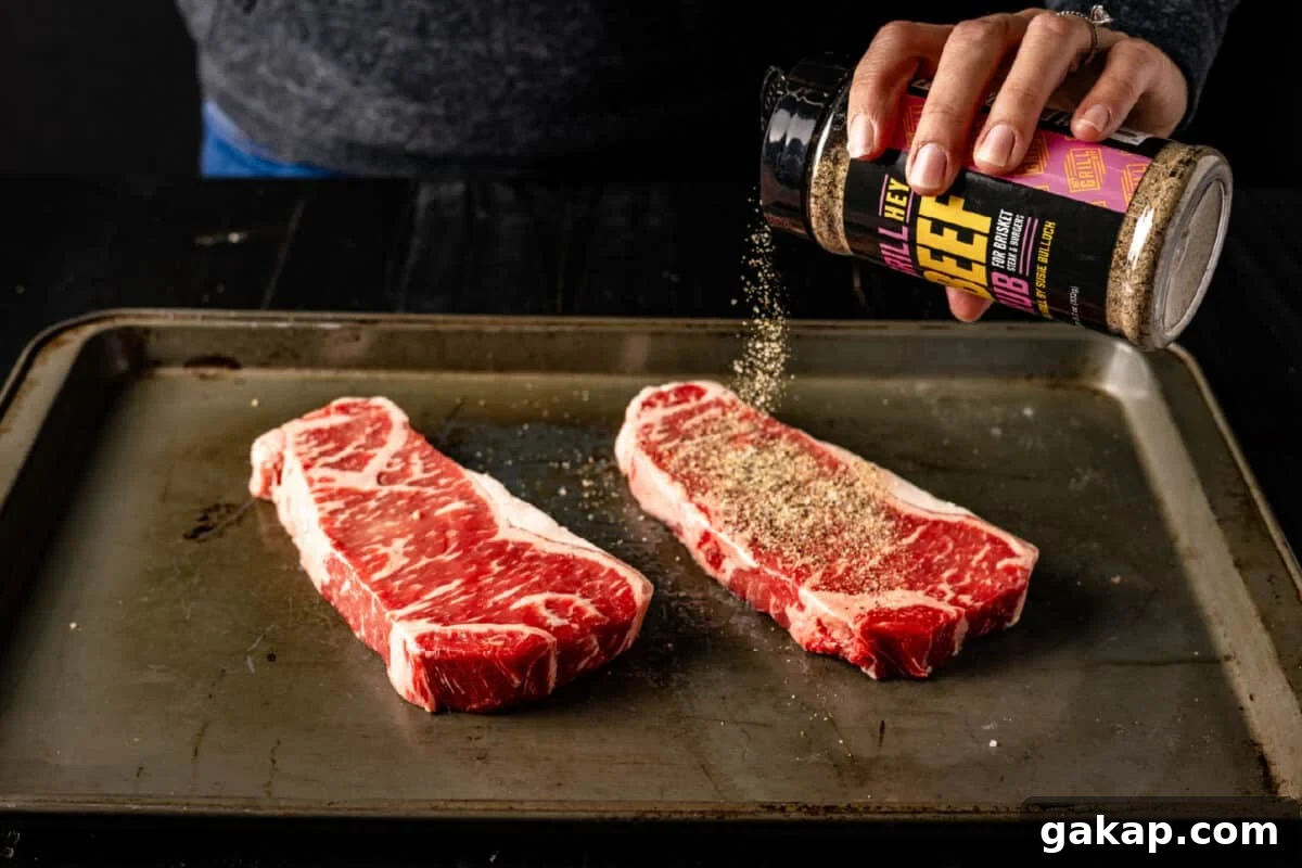 A chef's hand sprinkling Hey Grill Hey Beef Rub onto two uncooked steaks on a baking sheet.