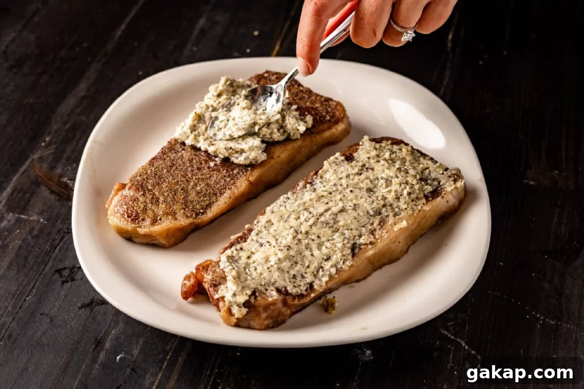 Two uncooked steaks on a white plate and a chef's hand using a spoon to spread butter onto one of the steak.