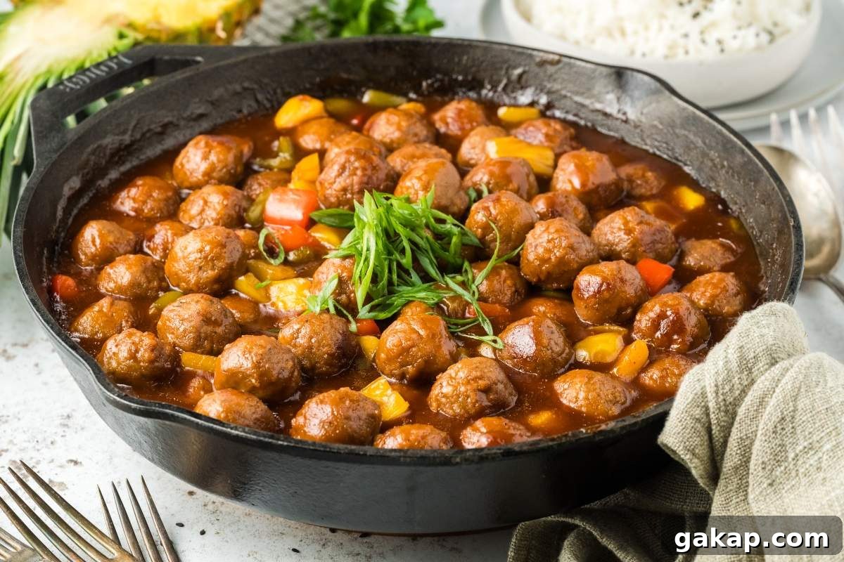 A cast-iron skillet brimming with sweet and sour meatballs garnished with green onions, placed alongside a bowl of rice and dining utensils.