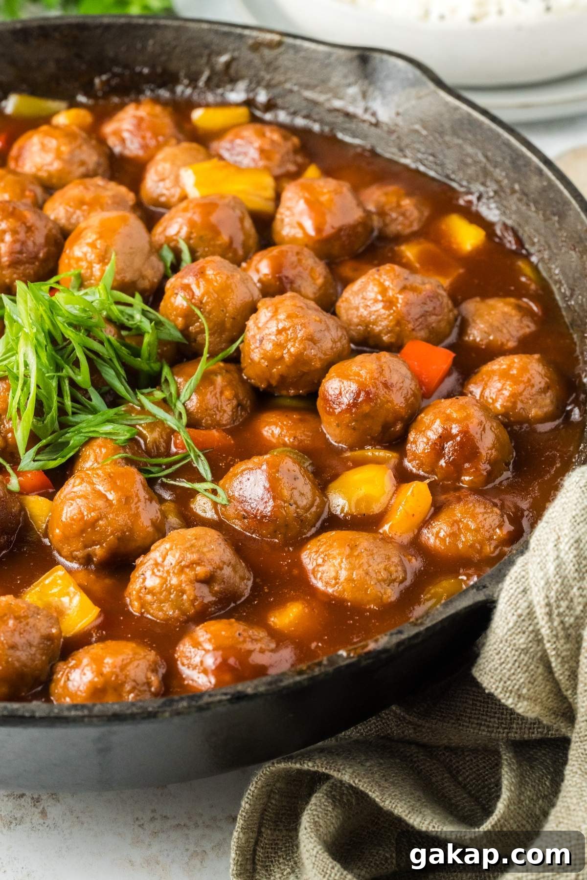 A skillet filled with sweet and sour meatballs in a rich sauce, topped with a garnish of chopped green onions and diced vegetables.