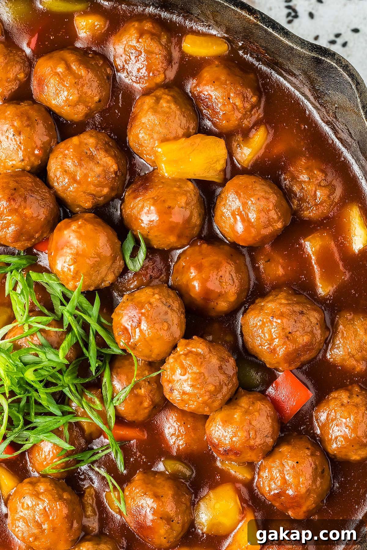Sweet and sour meatballs in a rich tomato sauce with diced red and yellow peppers, garnished with sliced green onions.