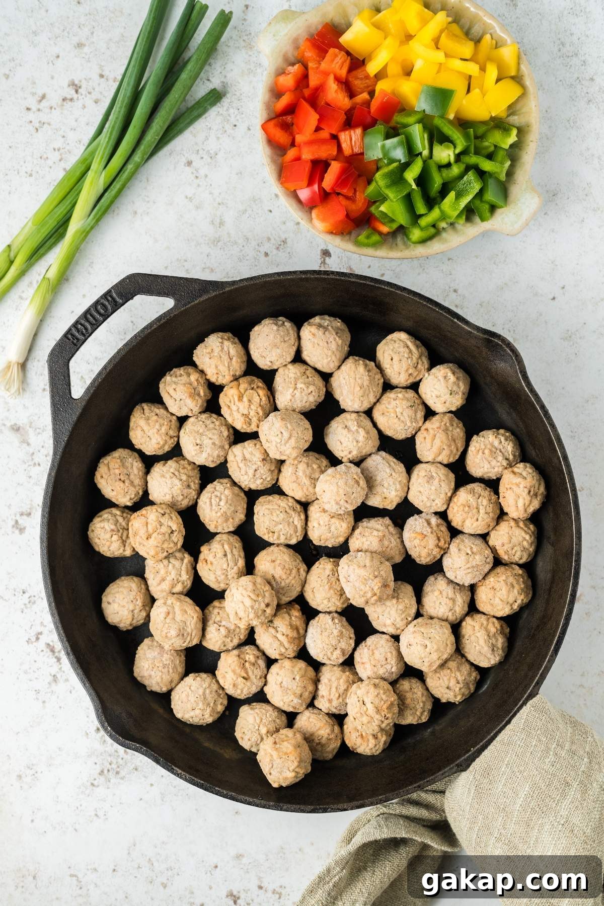 A cast iron skillet filled with uncooked sweet and sour meatballs sits next to a bowl of chopped red, yellow, and green bell peppers and chopped green onions.