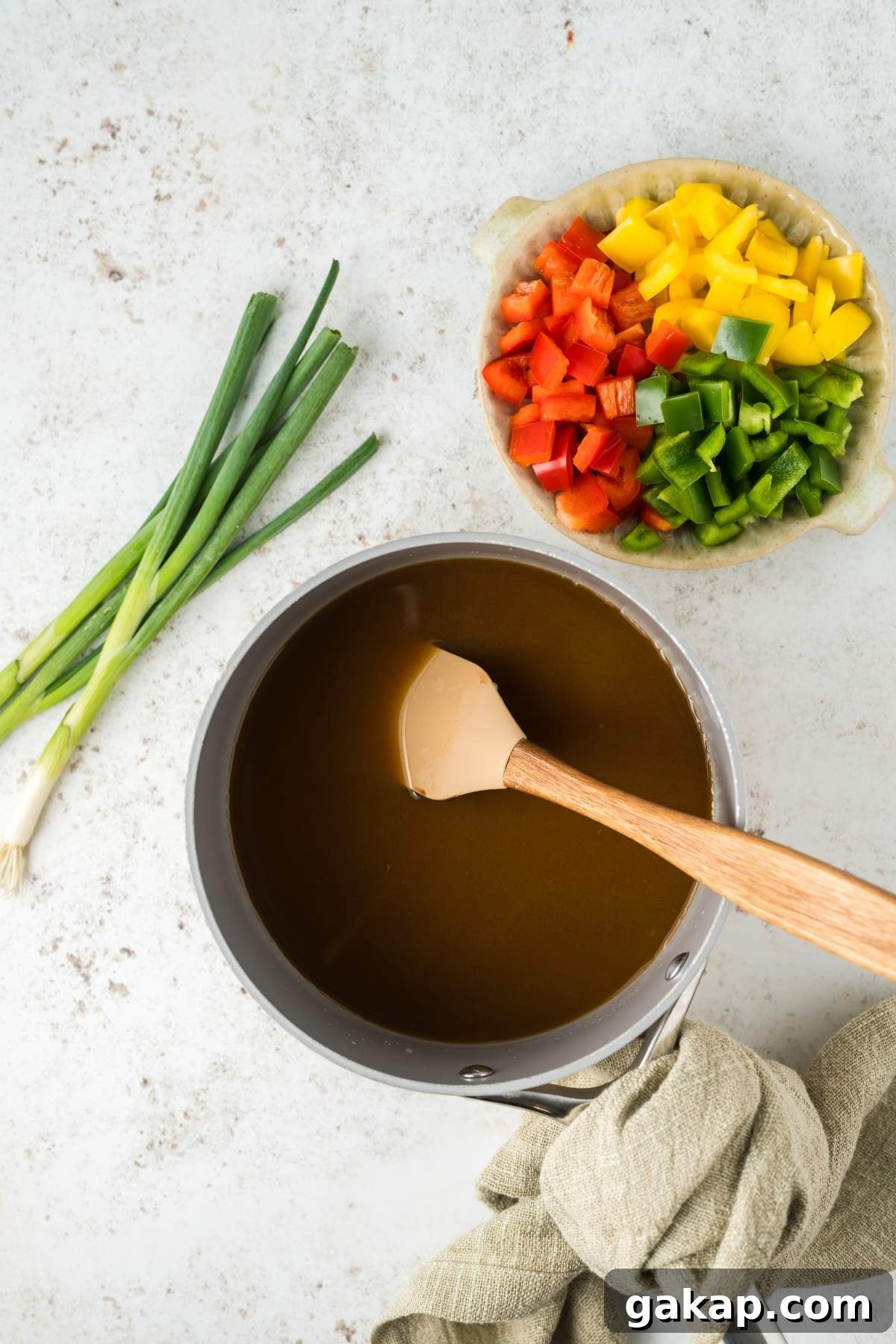 A saucepan filled with a dark liquid for sweet and sour meatballs, with a wooden spatula next to chopped red, yellow, and green bell peppers, and green onions on a light-colored surface.