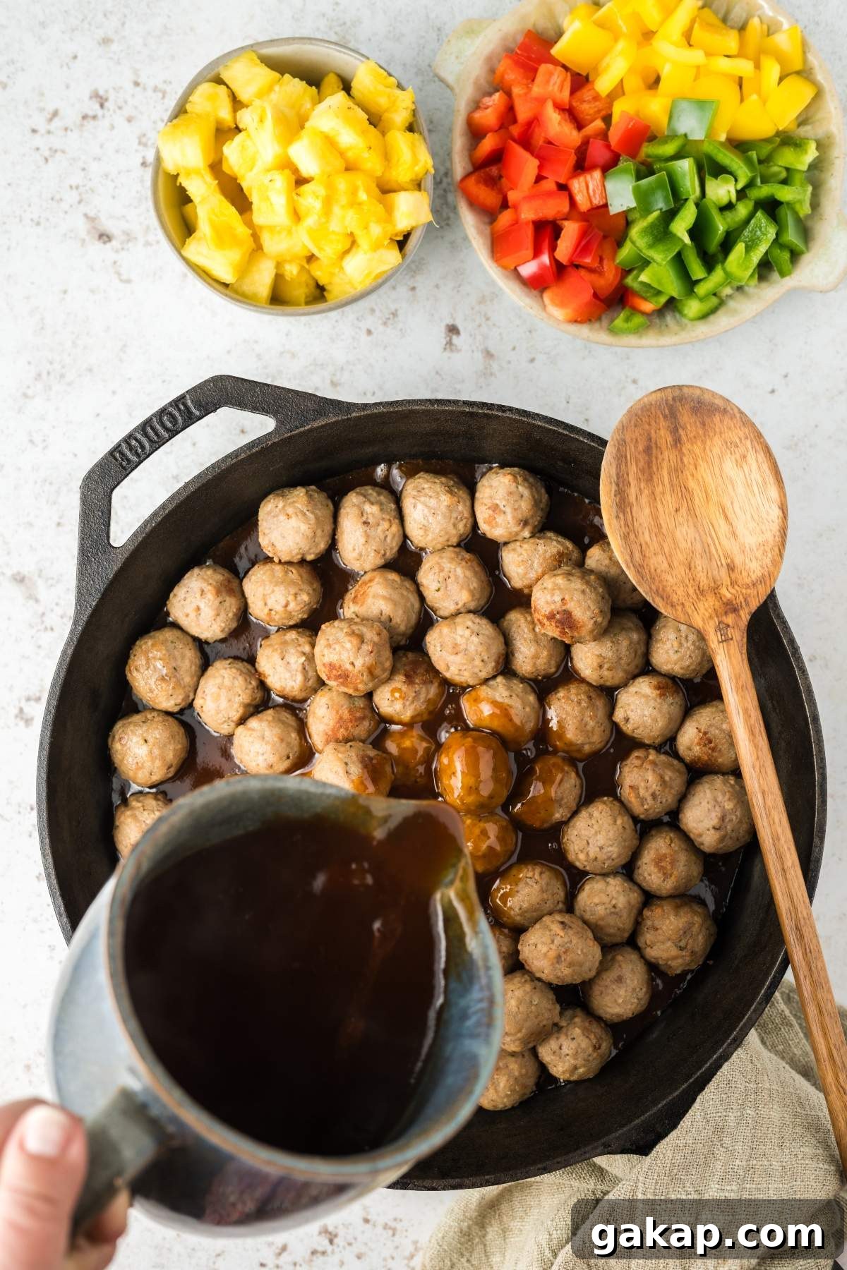 Top-down view of a skillet filled with sweet and sour meatballs, sauce being poured from a jug, and chopped pineapple, red, yellow, and green bell peppers in small bowls nearby. Wooden spoon on the side.