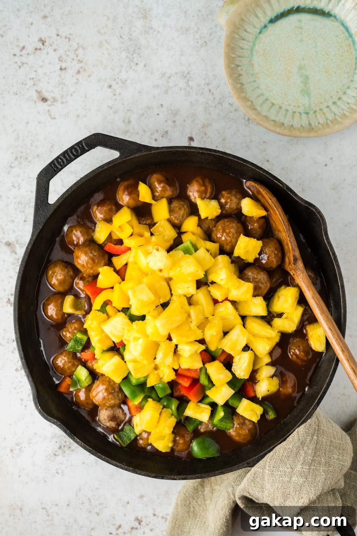 A skillet filled with sweet and sour meatballs, chopped pineapples, and bell peppers in a savory sauce, with a wooden spoon and a green napkin on the side. A small bowl is partially visible in the background.