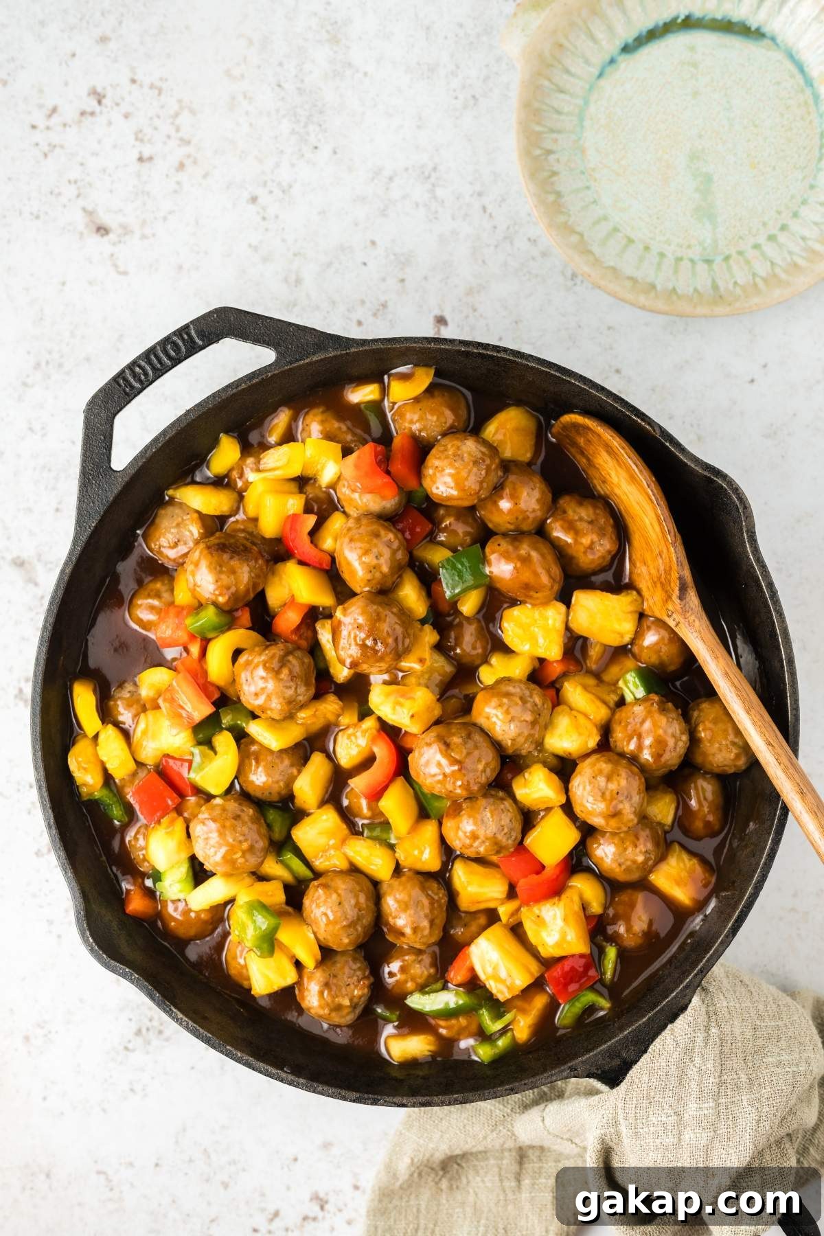A cast iron skillet with sweet and sour meatballs, bell peppers, and pineapple chunks in a sauce, with a wooden spoon resting inside it, is placed on a light-colored surface next to a cloth napkin and an empty bowl.