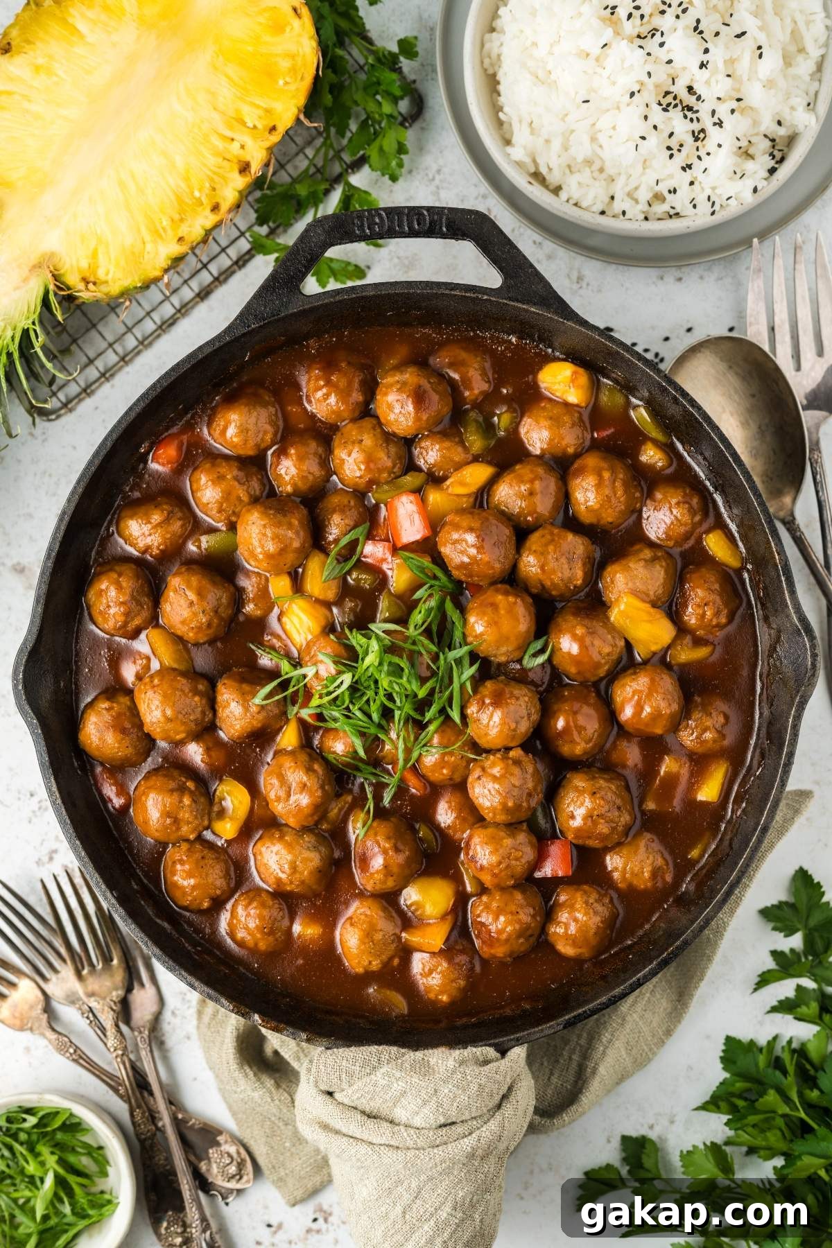 A cast iron skillet filled with sweet and sour meatballs in a rich, brown sauce, garnished with chopped green onions. The skillet is surrounded by fresh pineapple, white rice, and herbs on a white surface.