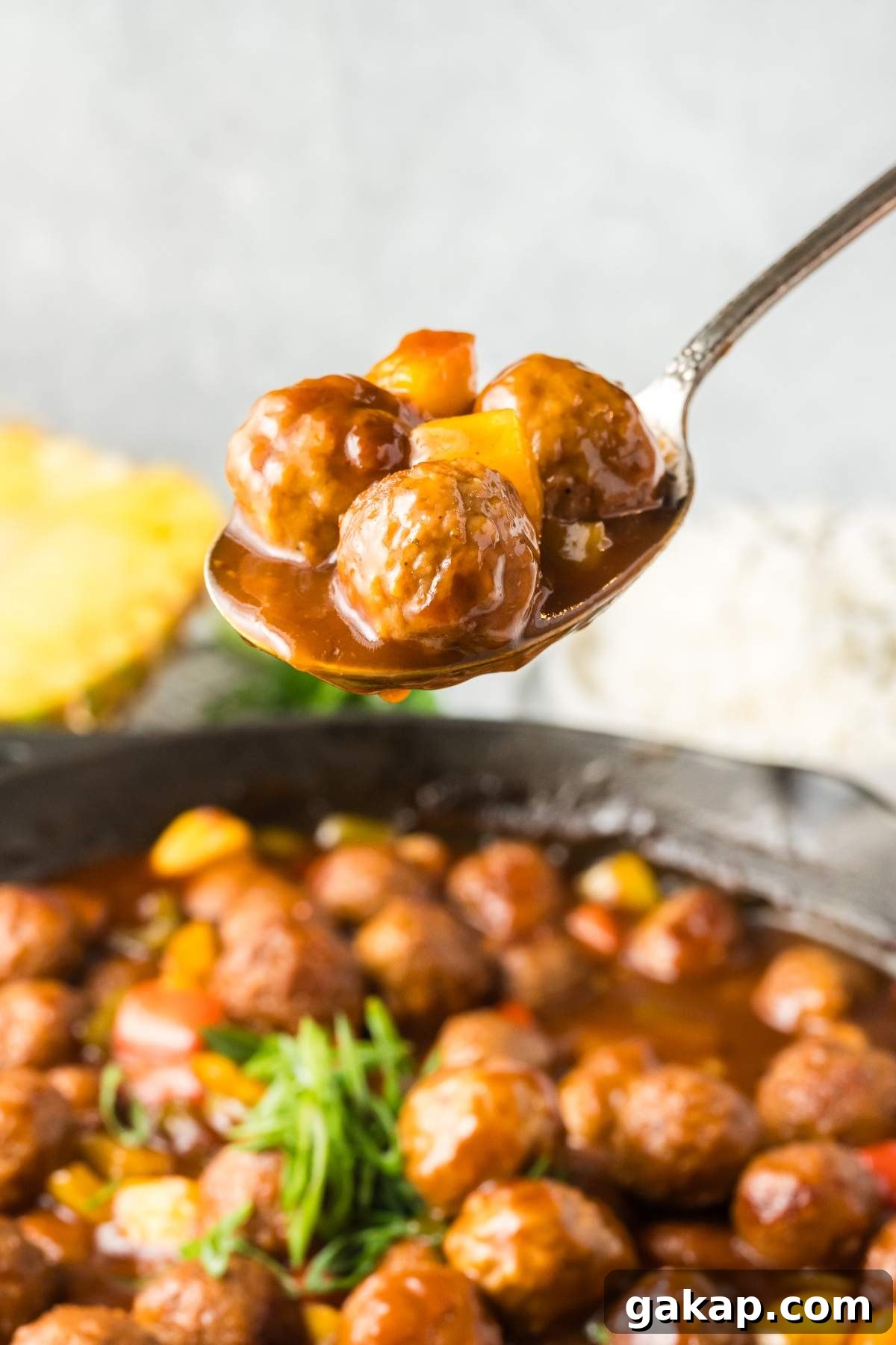 A spoonful of sweet and sour meatballs with diced vegetables is held above a skillet filled with more meatballs and sauce.