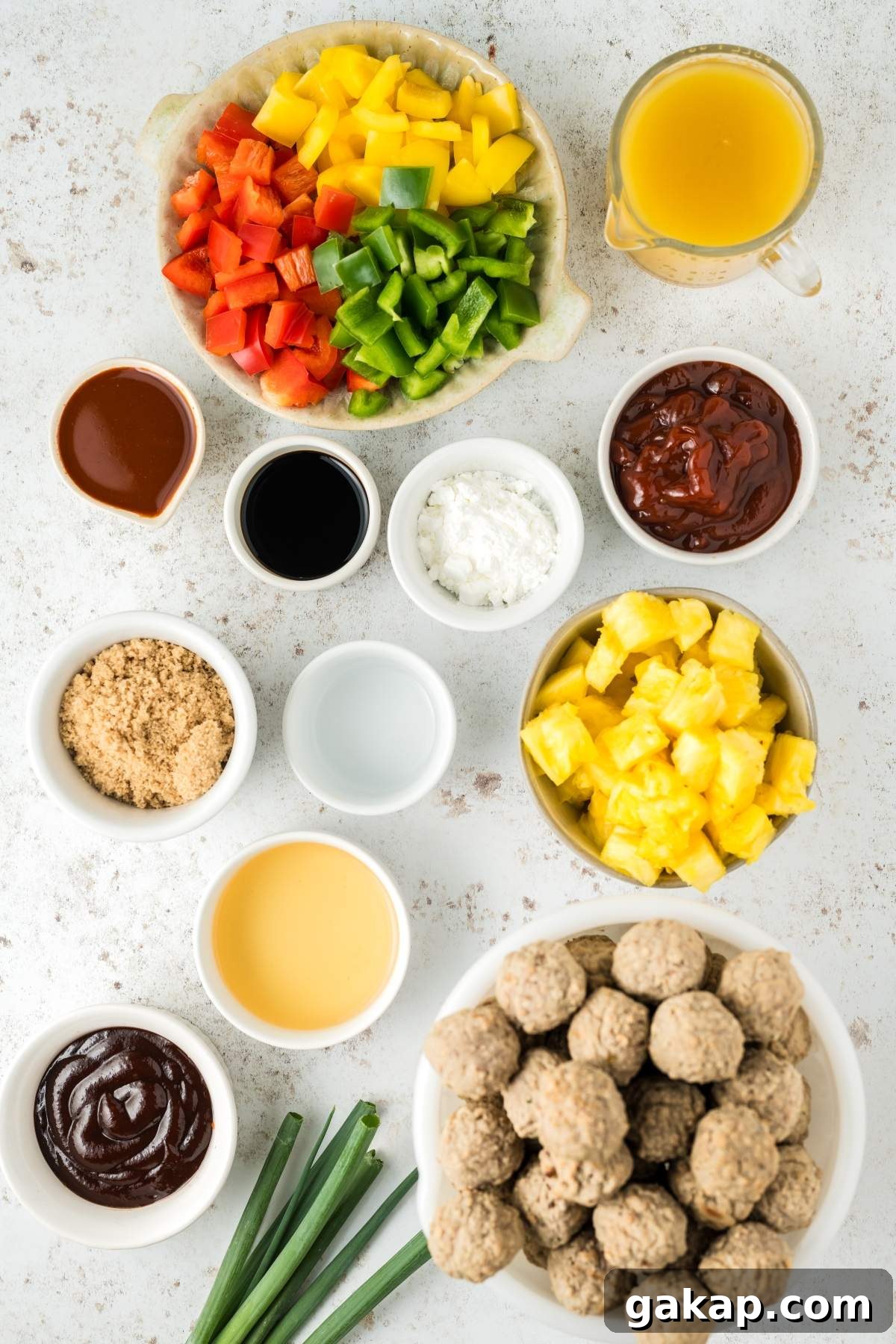 A variety of ingredients including chopped bell peppers, pineapple chunks, sweet and sour meatballs, sauces, brown sugar, cornstarch, and green onions are arranged in separate bowls on a countertop.