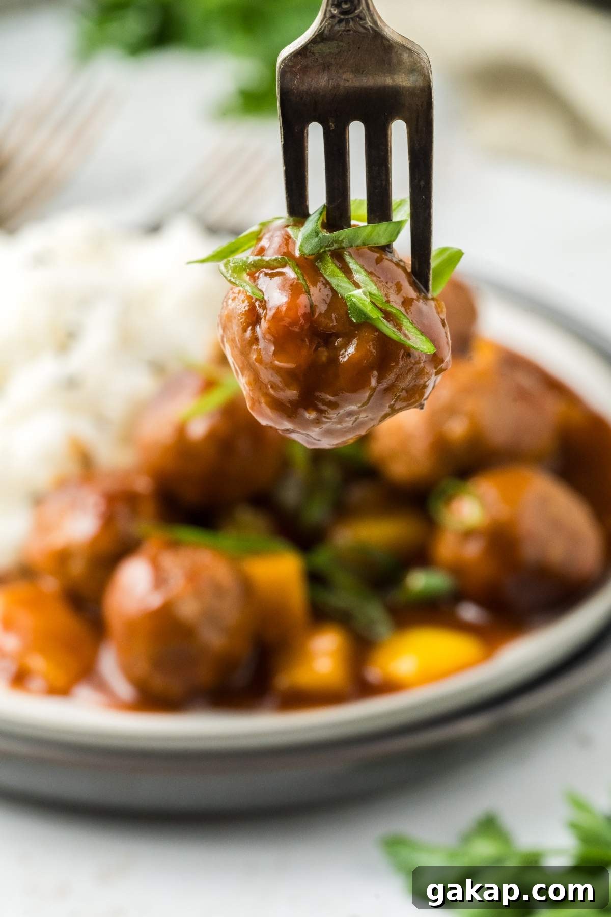 A fork holding a glazed sweet and sour meatball garnished with herbs, with a plate of more meatballs and a side of rice in the background.