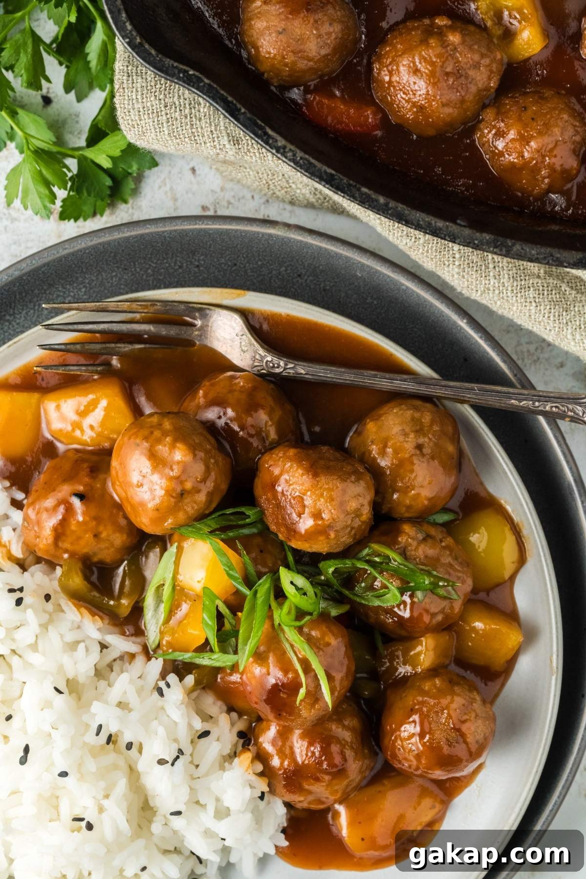 A plate with sweet and sour meatballs in a brown sauce, mixed with bell peppers, served alongside white rice garnished with black sesame seeds. A fork rests on the plate, and a skillet with more meatballs is in the background.
