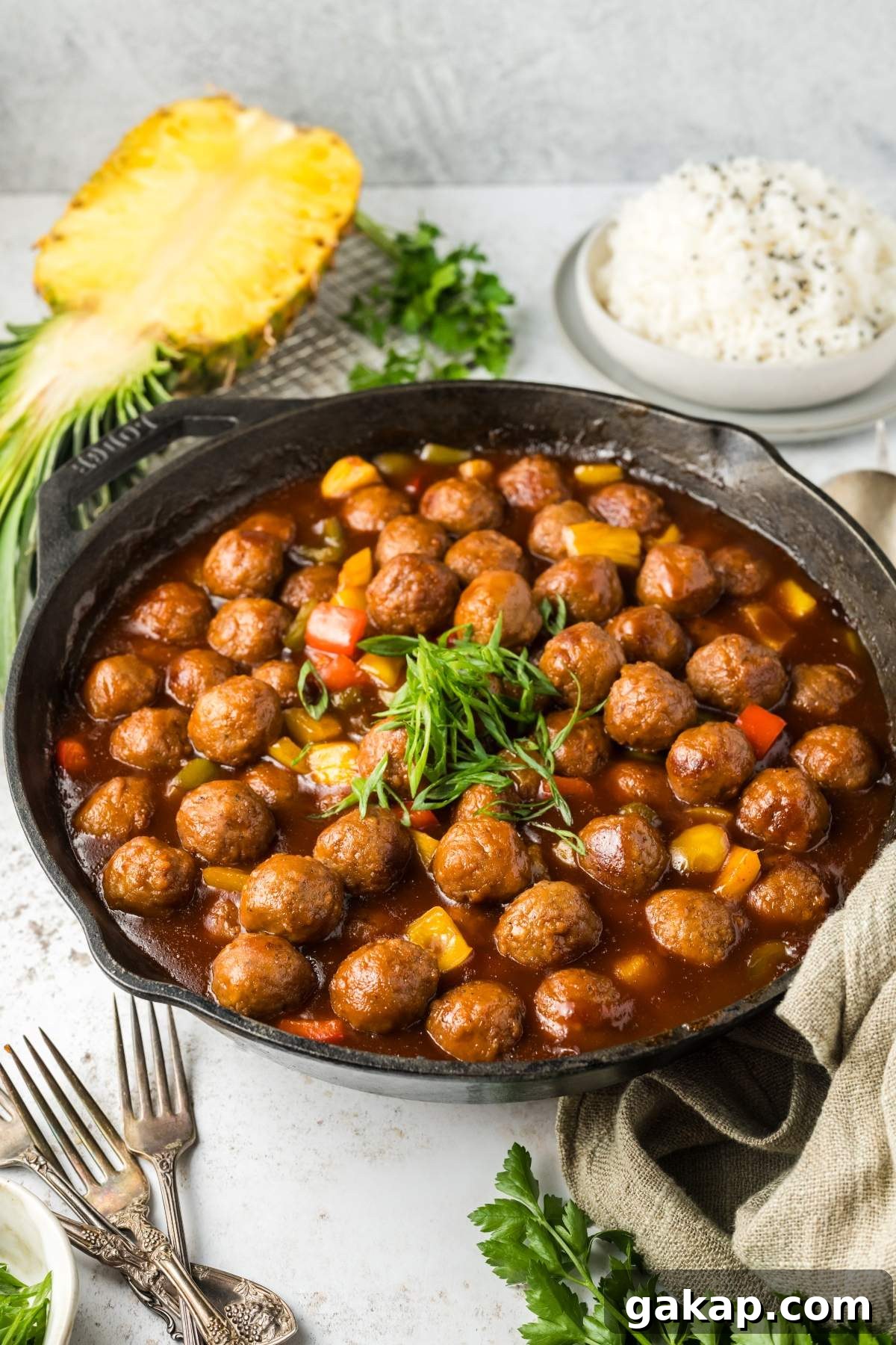A skillet filled with sweet and sour meatballs in a brown sauce, garnished with green onions, alongside a sliced pineapple, a bowl of white rice, and cutlery.