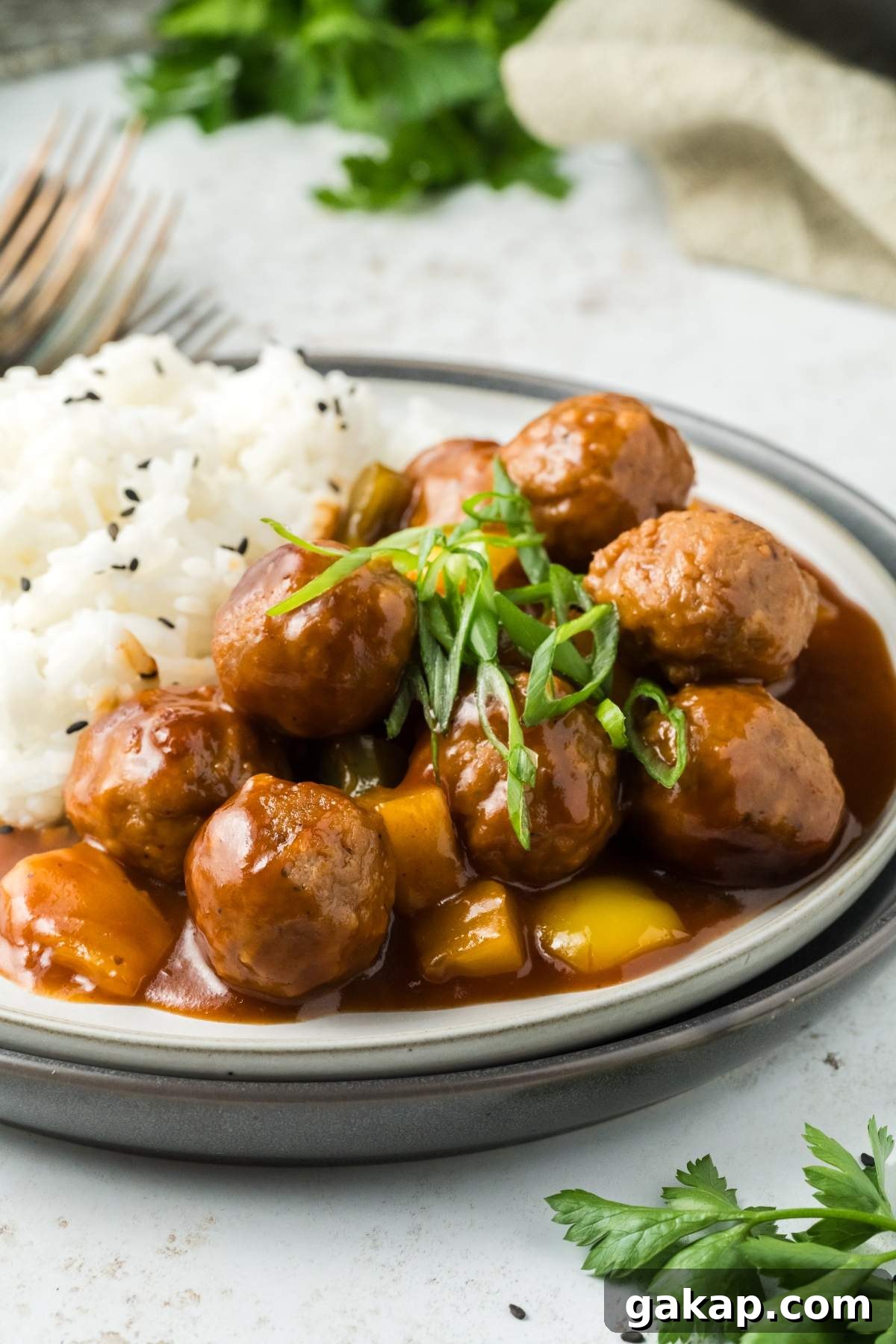 A plate of sweet and sour meatballs in a brown sauce with sliced green onions, served next to white rice with black sesame seeds on a white surface. Parsley and utensils are in the background.