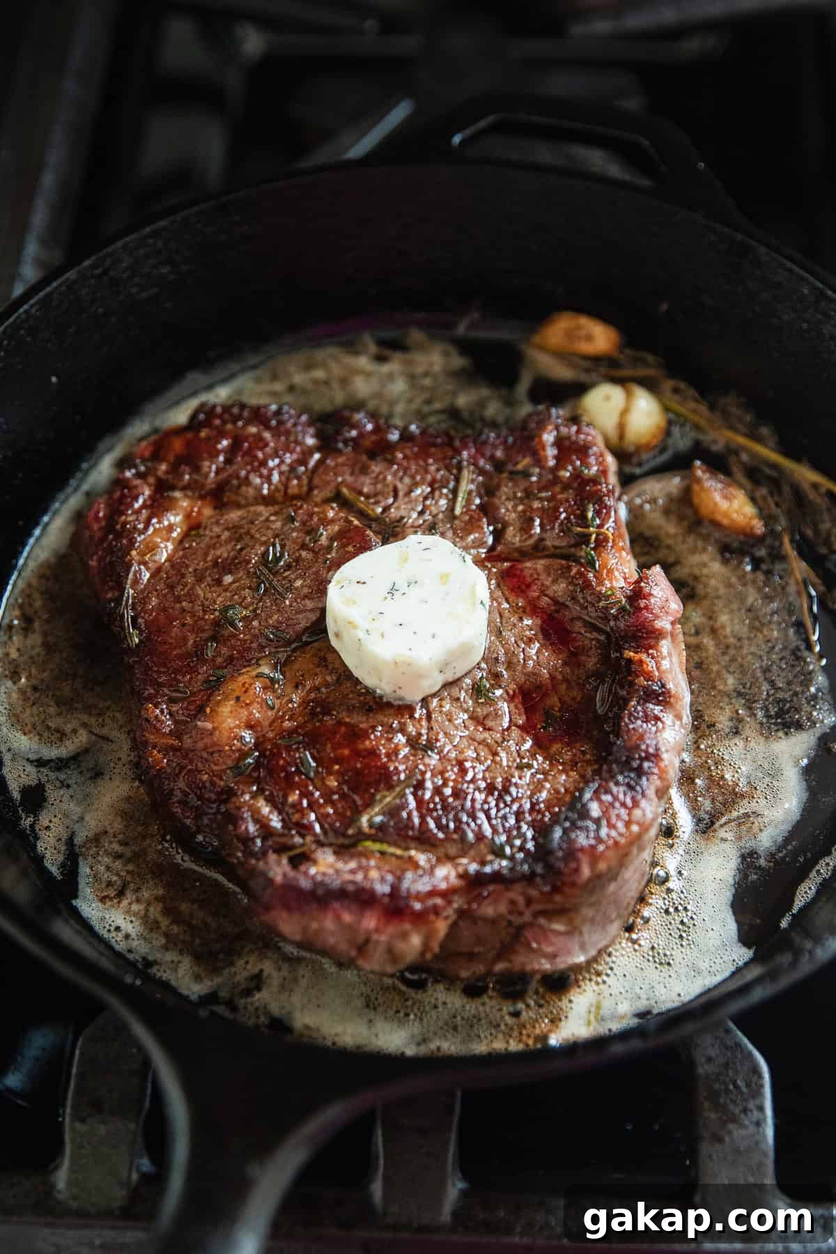 A ribeye steak cooking in a cast iron skillet with herb and garlic butter on top of steak.