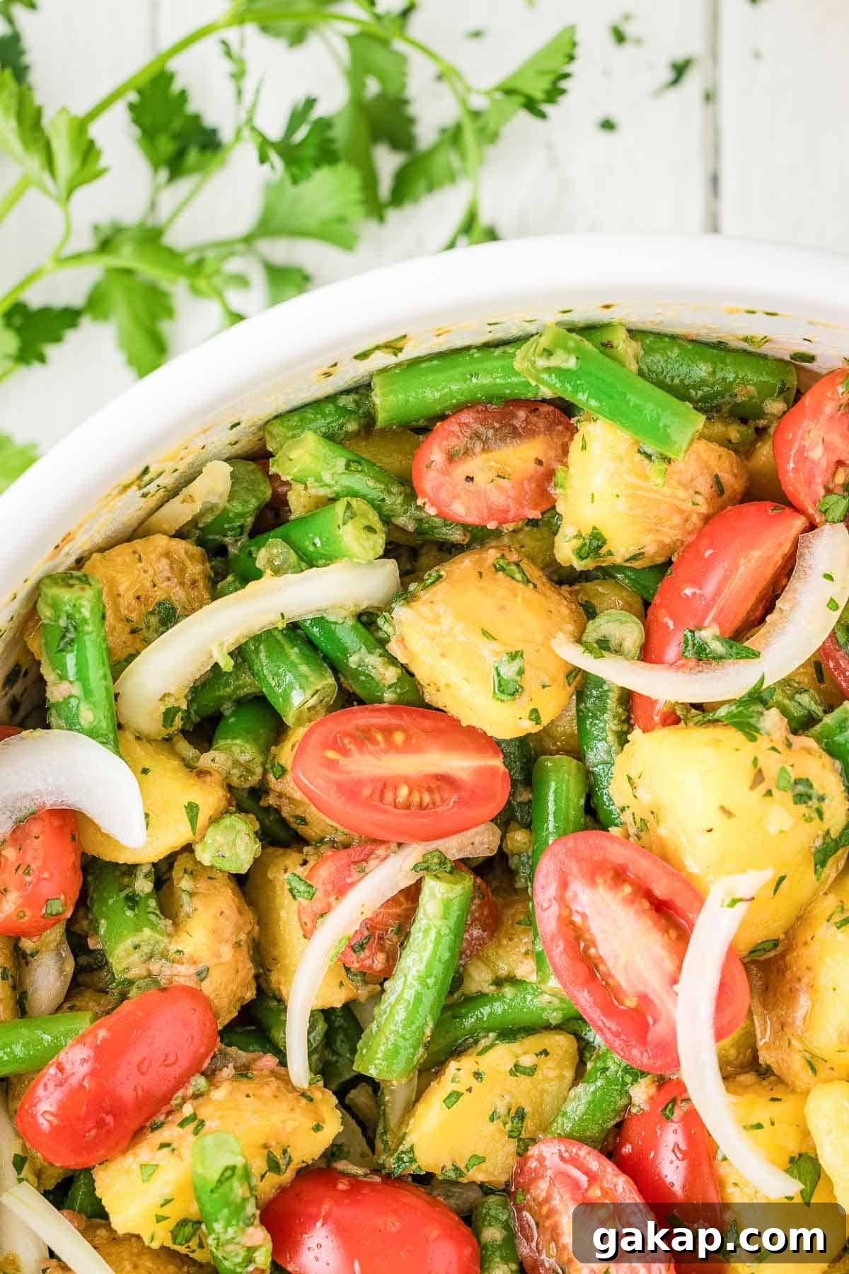 A bowl of potato salad with green beans, cherry tomatoes, sliced onions, and chopped herbs, with parsley scattered on a white wooden surface.