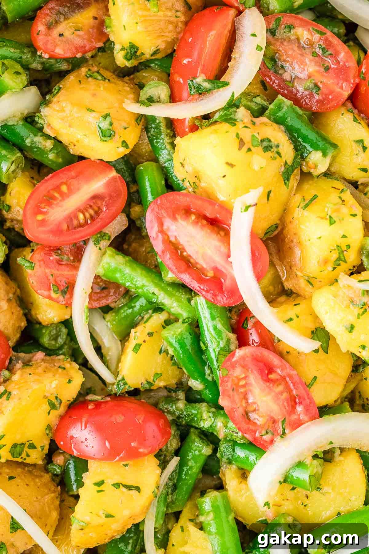 Close-up of a potato salad with halved cherry tomatoes, green beans, sliced onions, and herbs.
