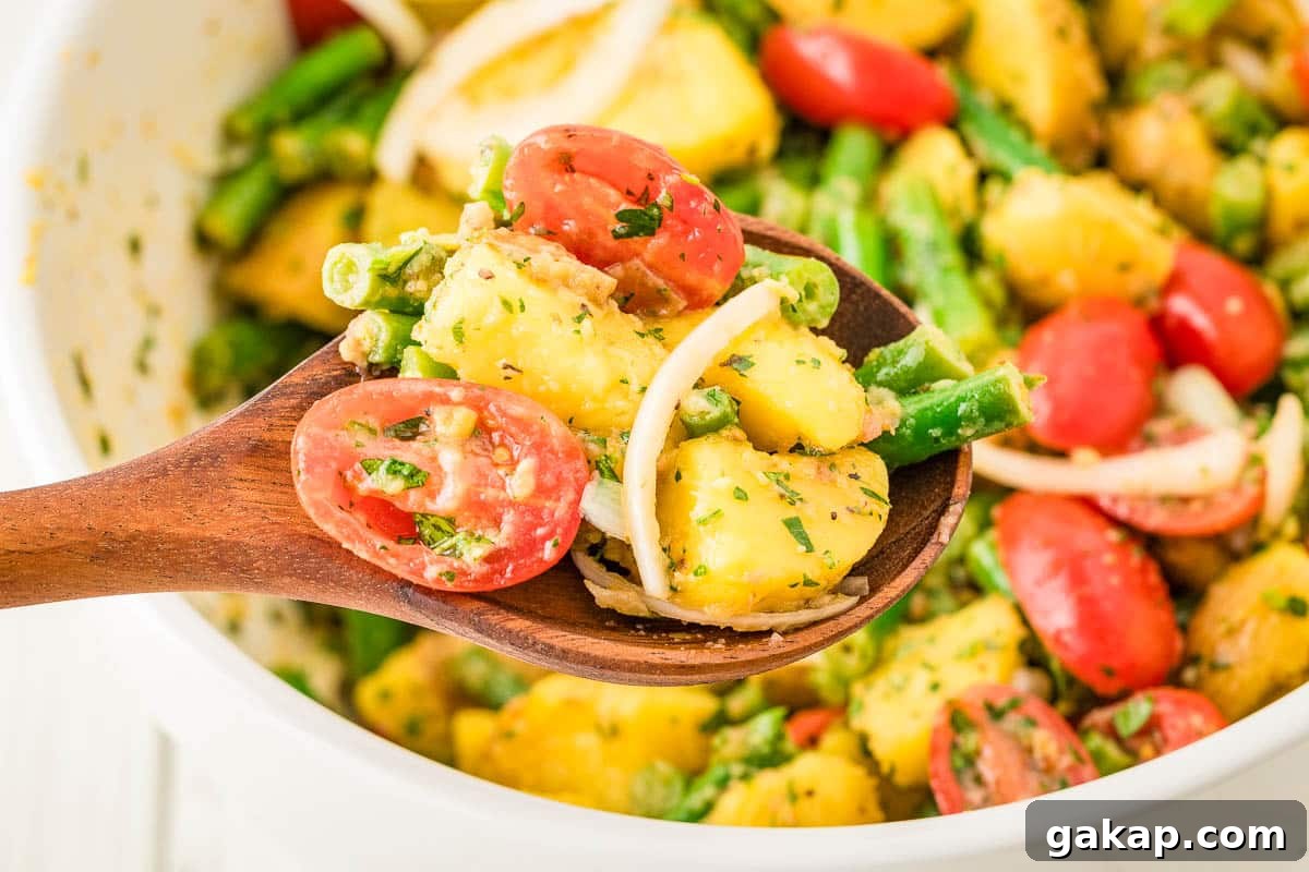 A wooden spoon holds a serving of potato salad with green beans, cherry tomatoes, onions, and herbs above a white bowl containing more of the salad.