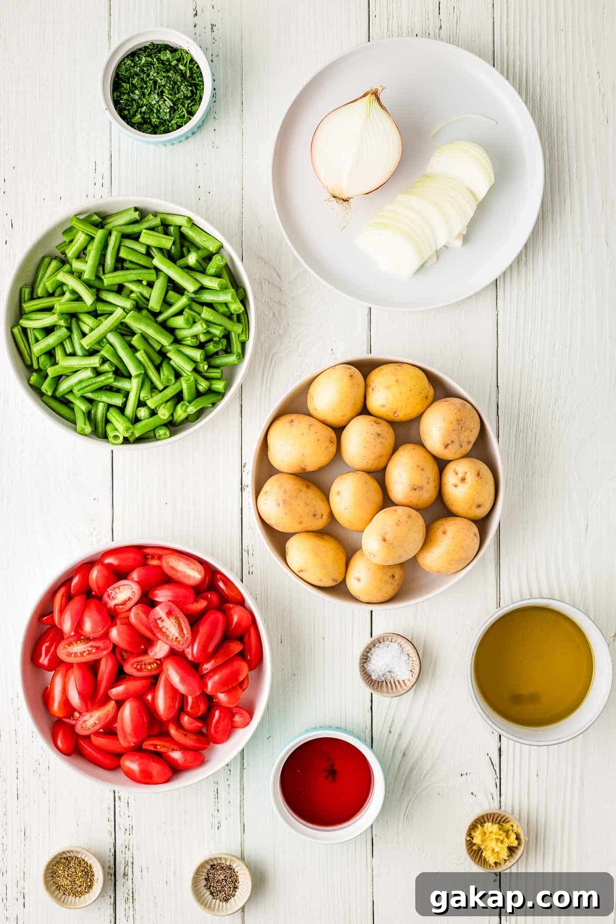 Bowls of halved cherry tomatoes, whole baby potatoes, chopped green beans, sliced onion, and various seasonings arranged on a white wooden surface.