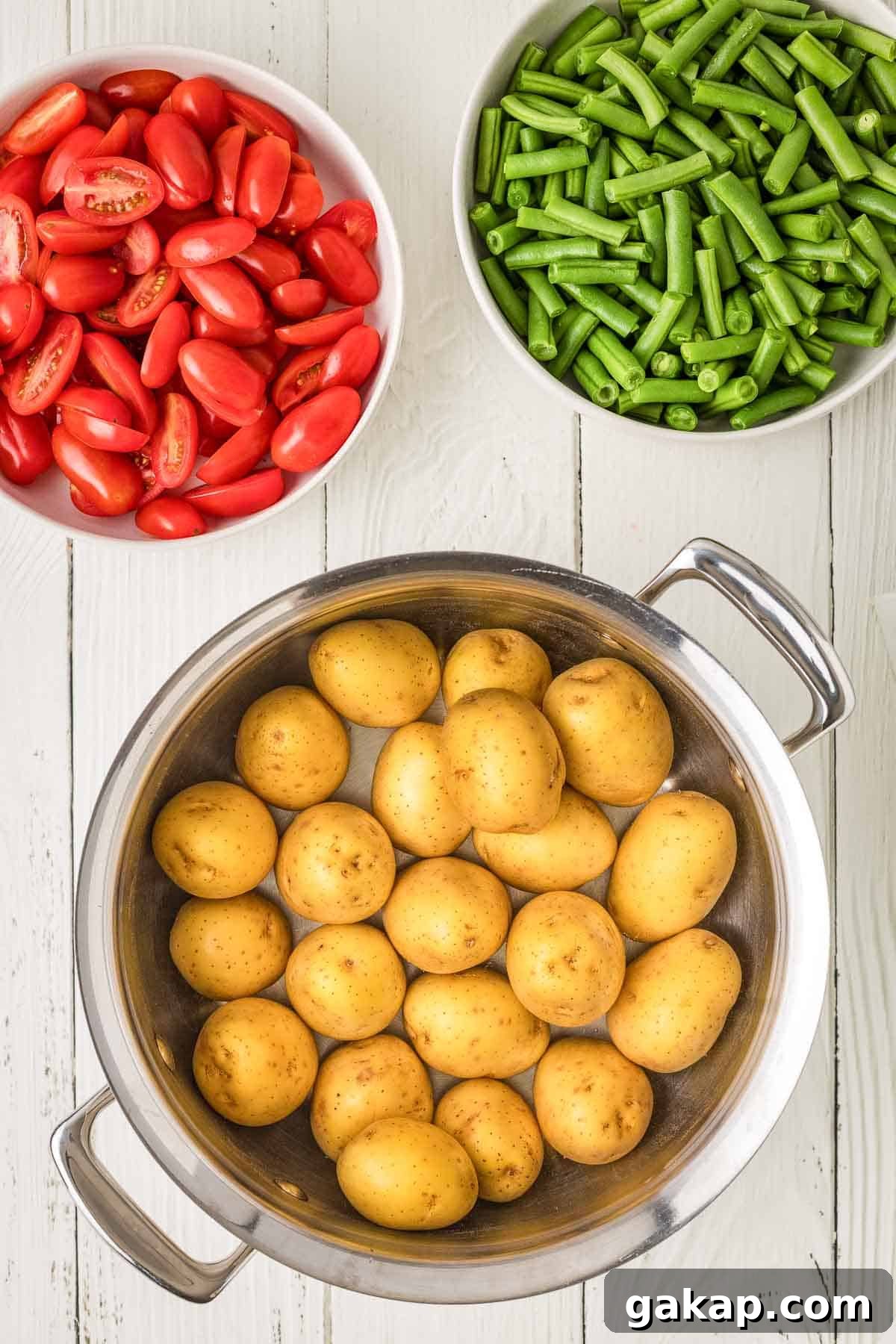 Three bowls on a white wooden surface: one with halved grape tomatoes, one with chopped green beans, and one with whole baby potatoes in a metal pot.