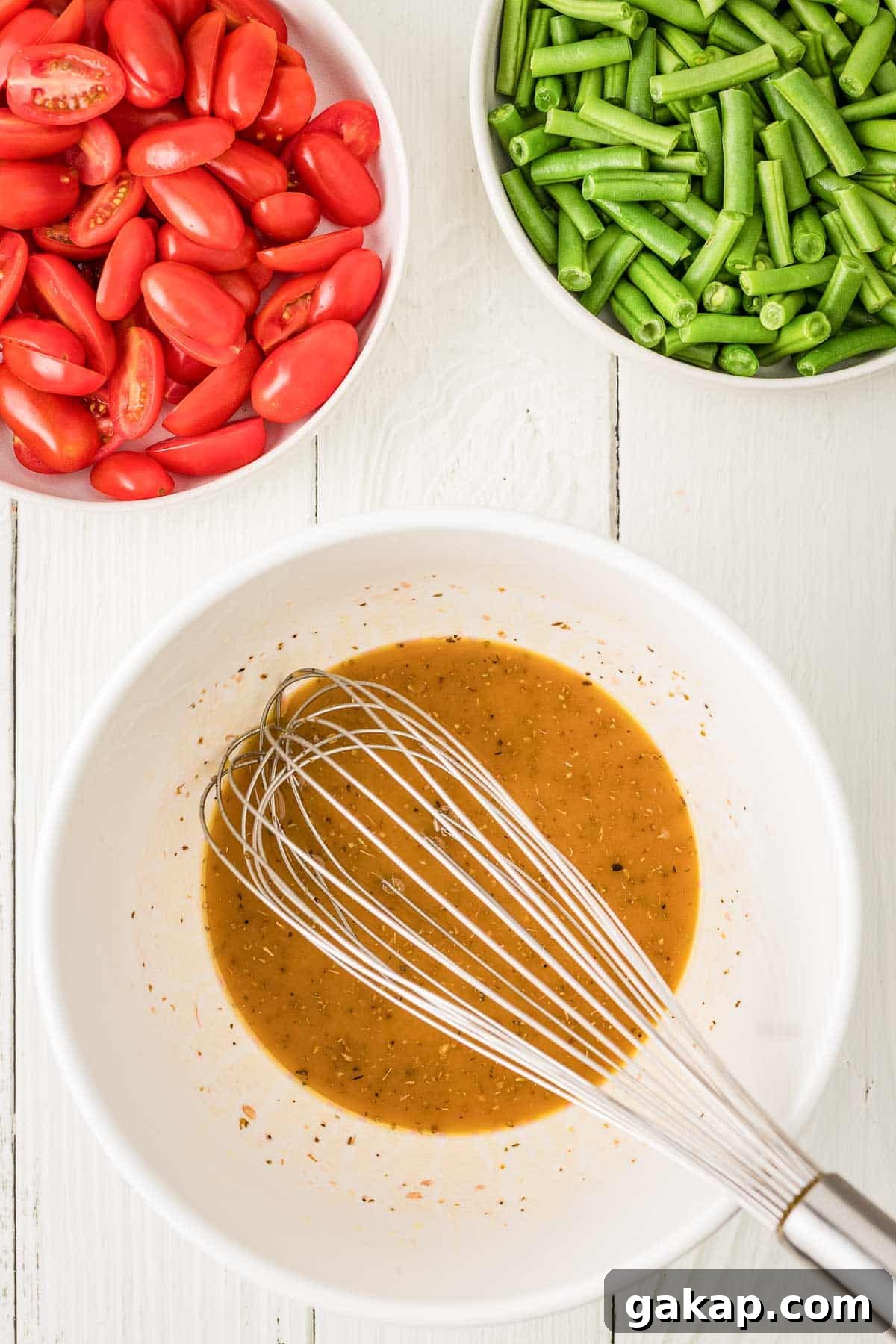 A metal whisk in a bowl of vinaigrette dressing, with bowls of chopped green beans and halved grape tomatoes on a white wooden surface.