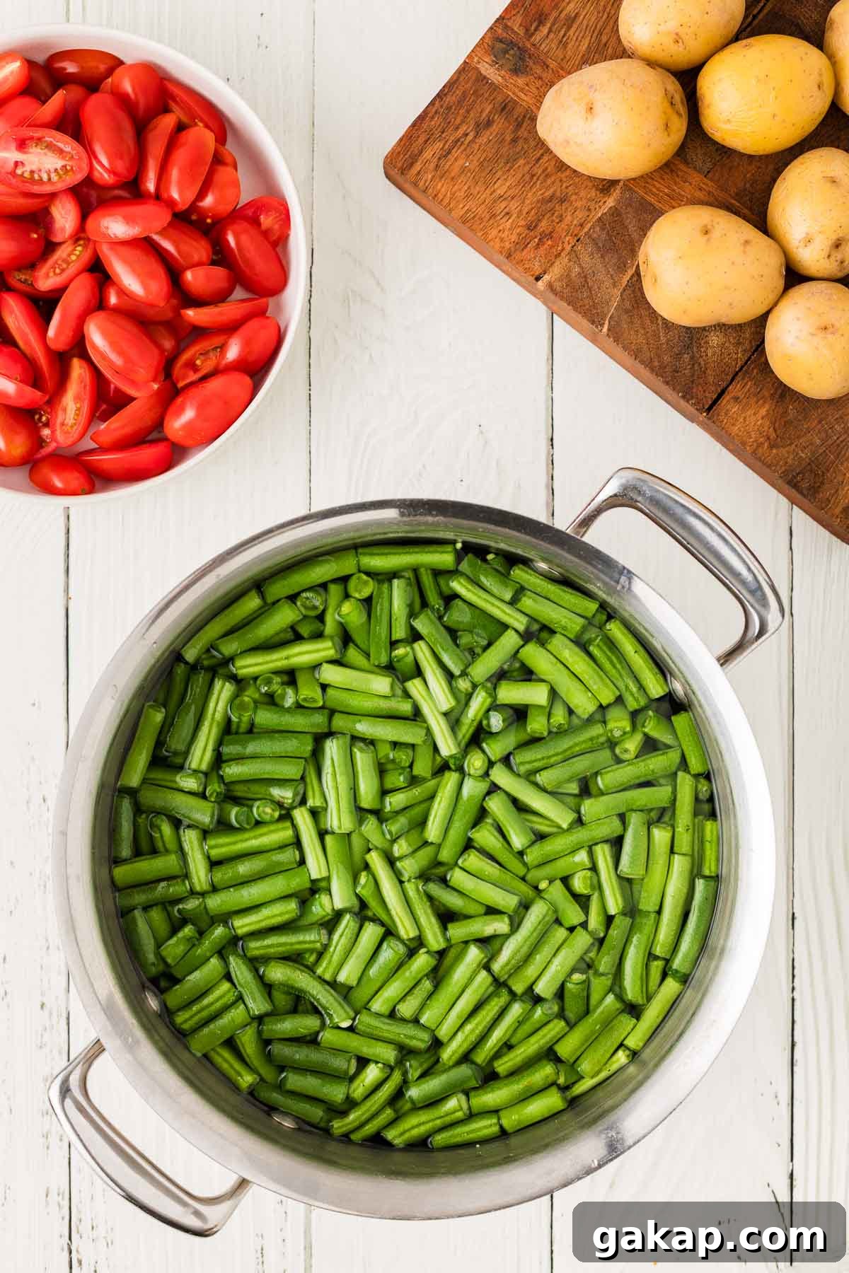A pot of cut green beans in water sits on a white wooden surface, next to a bowl of halved grape tomatoes and a cutting board with whole yellow potatoes.