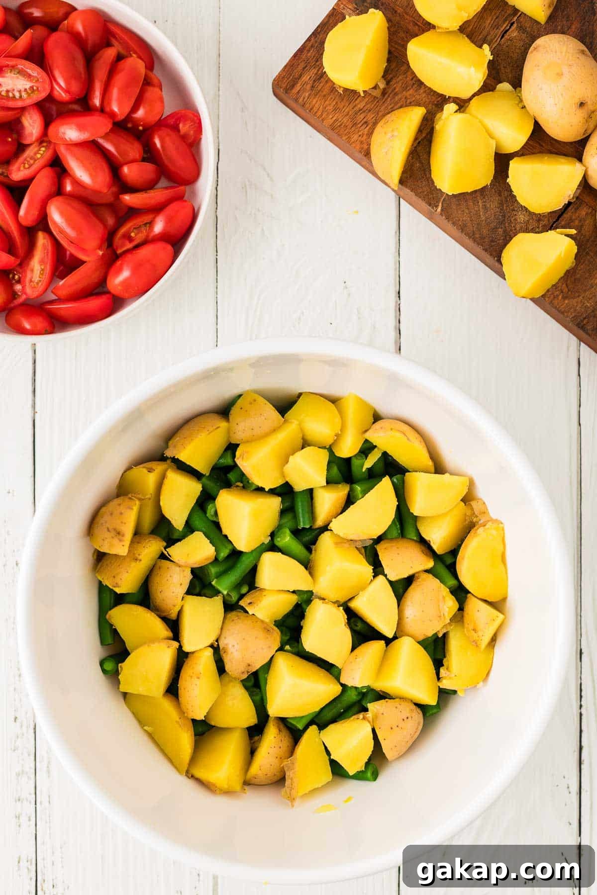 A white bowl with chopped yellow potatoes and green beans, a cutting board with more potatoes, and a bowl of halved cherry tomatoes on a white wooden surface.