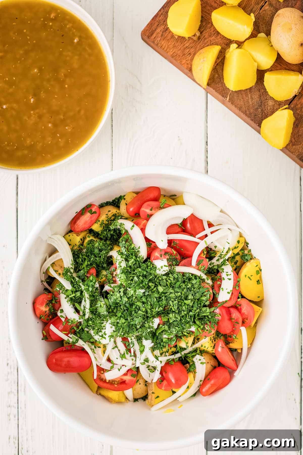 A white bowl filled with sliced tomatoes, onions, potatoes, and chopped herbs sits on a white table; a cutting board with potatoes and a bowl of broth are nearby.