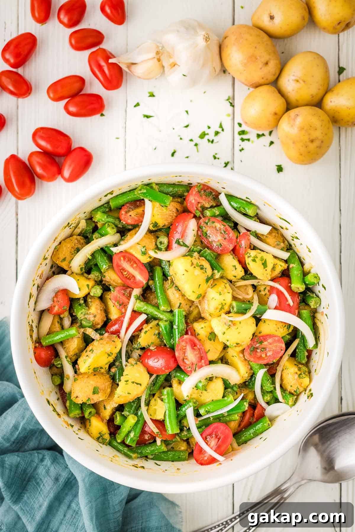 A bowl of potato salad with green beans, cherry tomatoes, onions, and herbs sits on a white table, surrounded by fresh potatoes, garlic, and tomatoes.