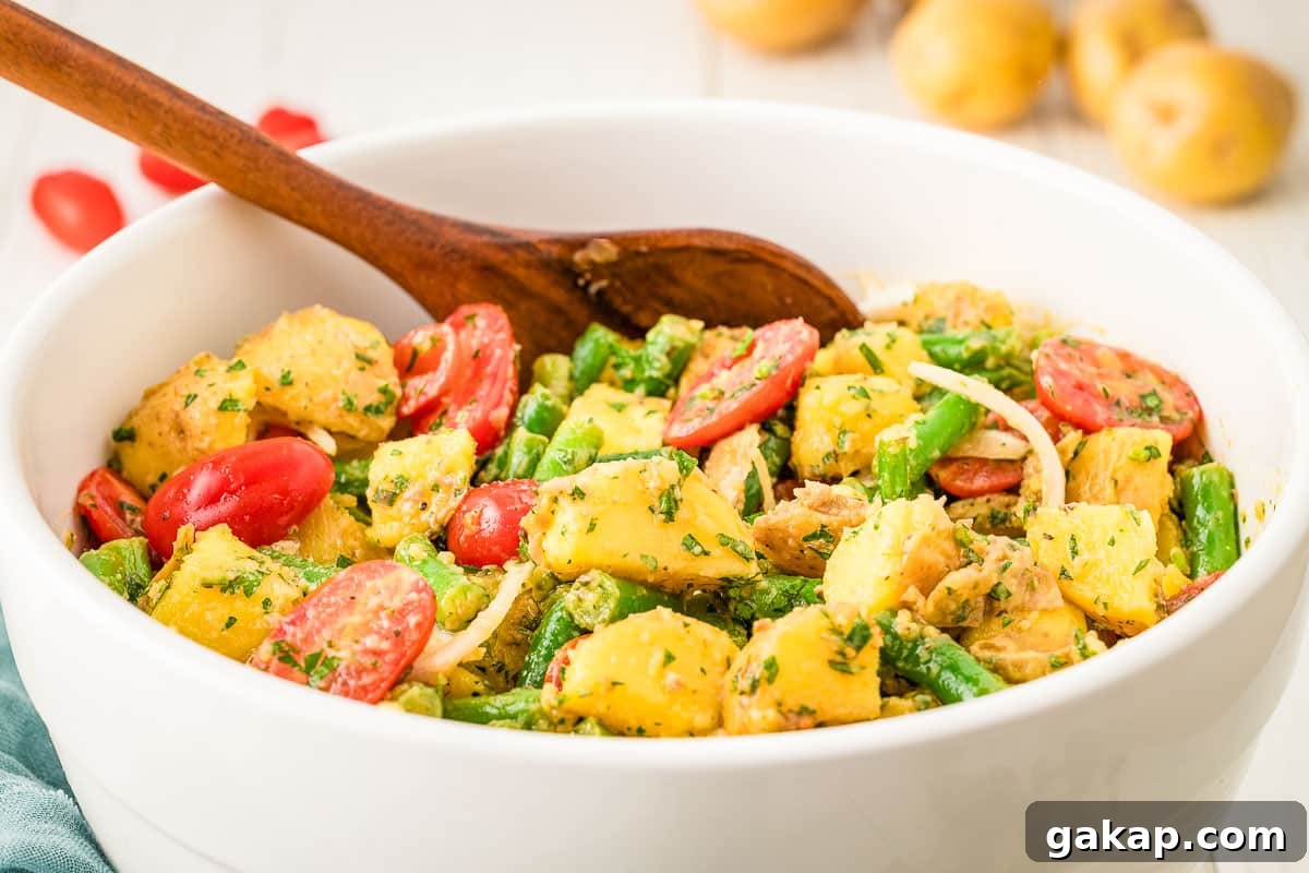 A white bowl filled with potato salad, green beans, cherry tomatoes, onions, and herbs, with a wooden spoon resting inside.
