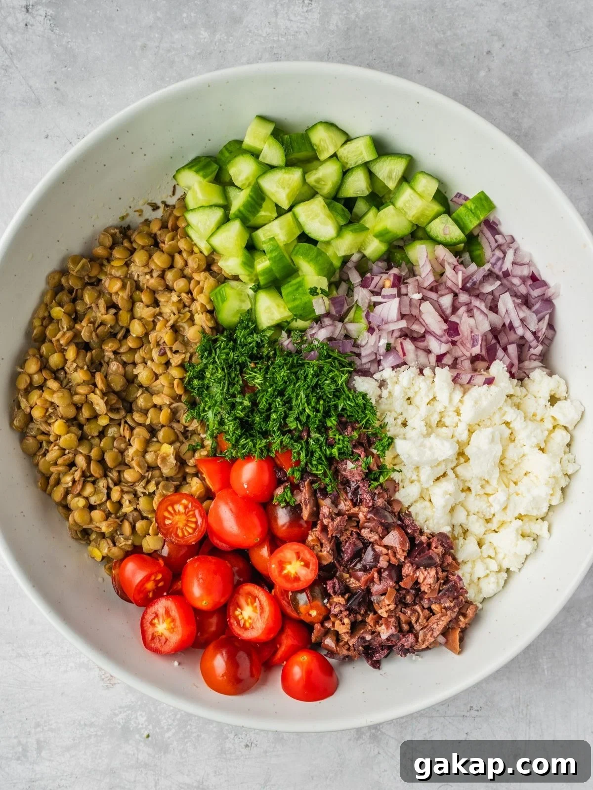 Large bowl with cooked lentils, chopped cucumbers, sliced cherry tomatoes, crumbled feta, chopped olives and herbs.
