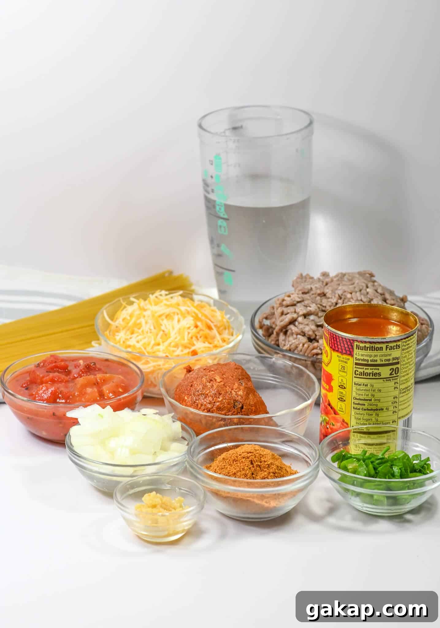 Craft the perfect dish with pasta, canned tomatoes, shredded cheese, ground meat, onions, green onions, garlic, and seasonings. A refreshing glass of water awaits in the background to complement this flavorful dish.