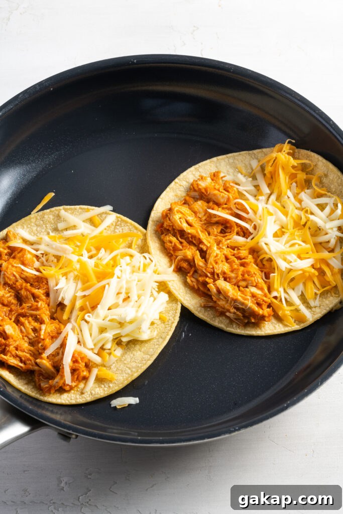 two corn tortillas topped with mixed shredded cheese on the right halves and saucy chicken tinga on the left halves in a skillet with oil.