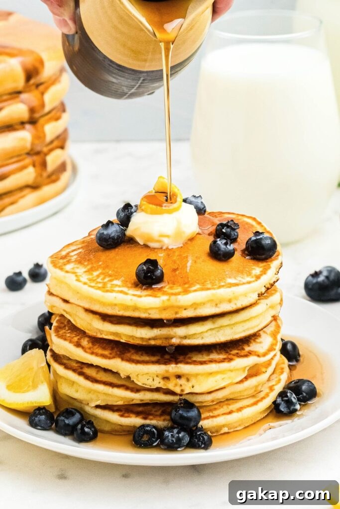 Lemon Pancakes Recipe: Fluffy Citrus Pancakes with Zesty Syrup 2 Golden fluffy lemon pancakes stacked on a white plate with syrup being poured on top.