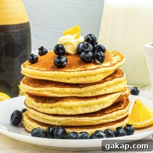 Lemon Pancakes Recipe: Fluffy Citrus Pancakes with Zesty Syrup 12 Golden fluffy pancakes stacked on a white plate, topped with blueberries and butter.