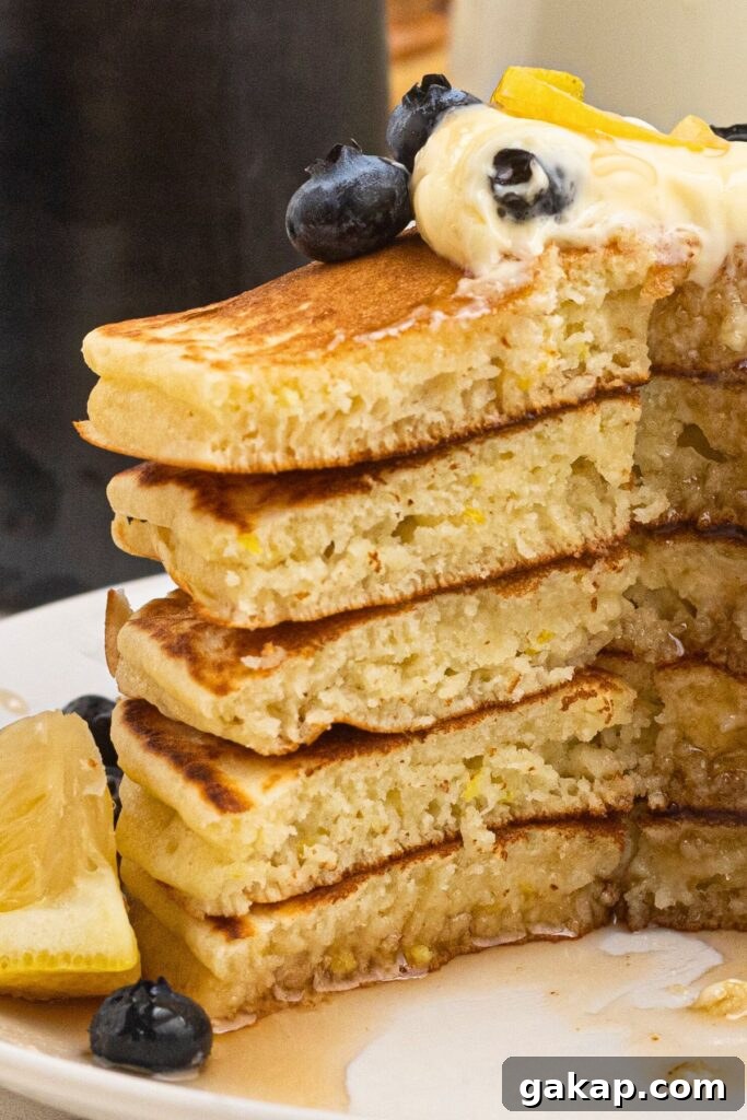 Lemon Pancakes Recipe: Fluffy Citrus Pancakes with Zesty Syrup 7 Light and fluffy pancakes stacked on a white plate, drizzled in syrup with butter and blueberries on top.