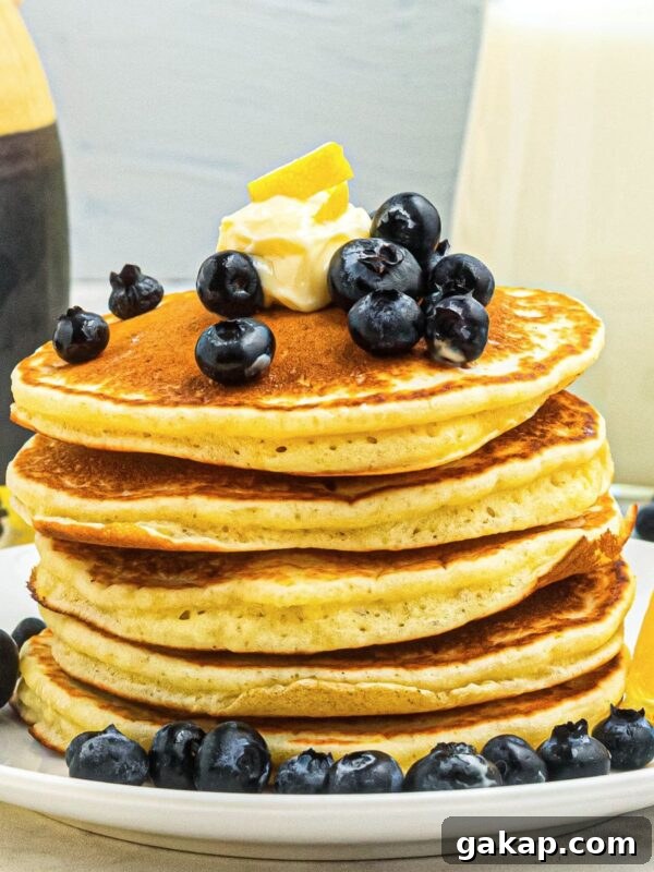 Lemon Pancakes Recipe: Fluffy Citrus Pancakes with Zesty Syrup 8 Golden fluffy pancakes stacked on a white plate, topped with blueberries and butter.