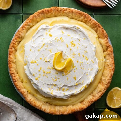 Lemon cream pie topped with fresh whipped cream and lemon zest centered on a green tiled background surrounded by lemons, wood dessert plates, forks, napkin, and pie server.