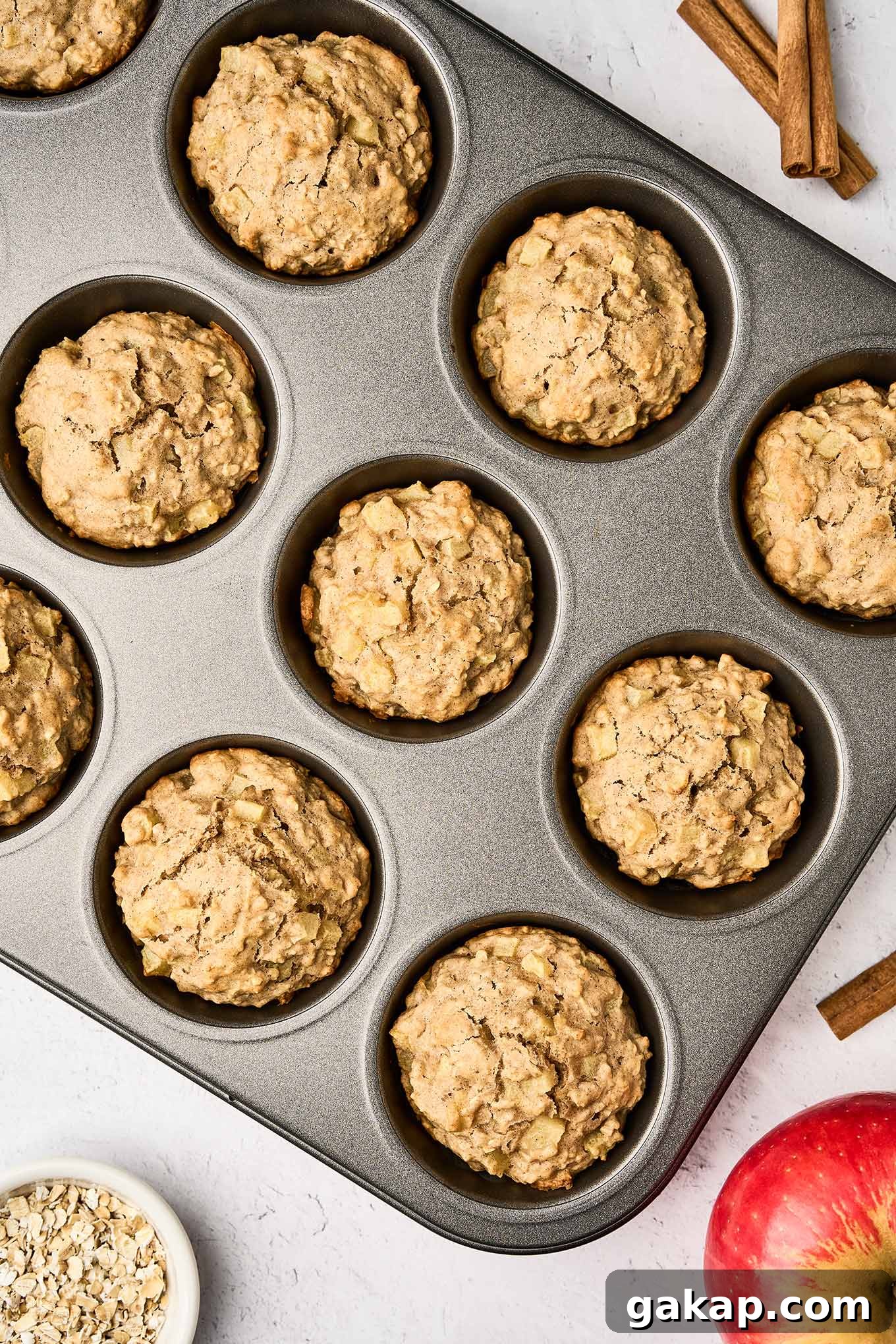 Buttermilk Apple Oat Muffins Recipe for Moist Breakfast 6 An overhead view of apple oatmeal muffins fresh from the oven in the muffin tin, surrounded by a whole apple, cinnamon sticks, and a small bowl of oats.