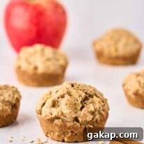 Buttermilk Apple Oat Muffins Recipe for Moist Breakfast 7 A single apple oatmeal muffin with more muffins and a whole apple in the background, cinnamon sticks and scattered oats in the foreground.