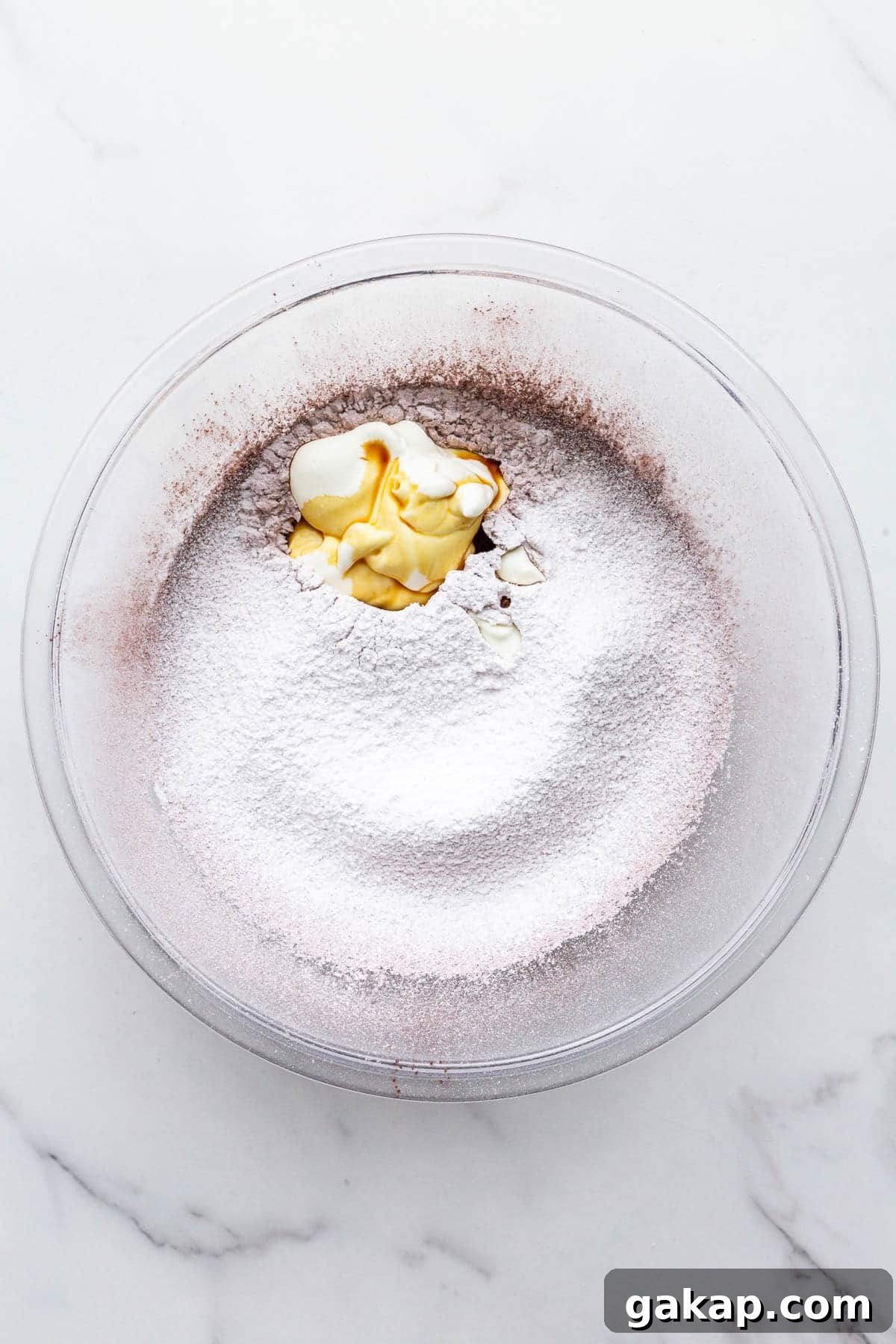Adding sour cream and vanilla to a glass bowl with sifted icing sugar and cocoa powder to make a chocolate fudge frosting.