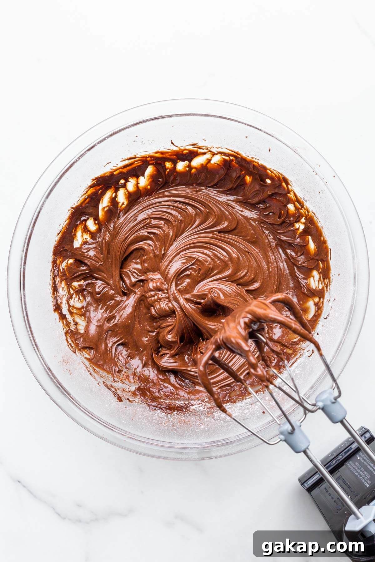 A glass bowl of glossy chocolate fudge frosting mixed with an electric mixer.