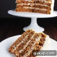 Slice of four layer carrot cake on a plate with cake on a stand in background.