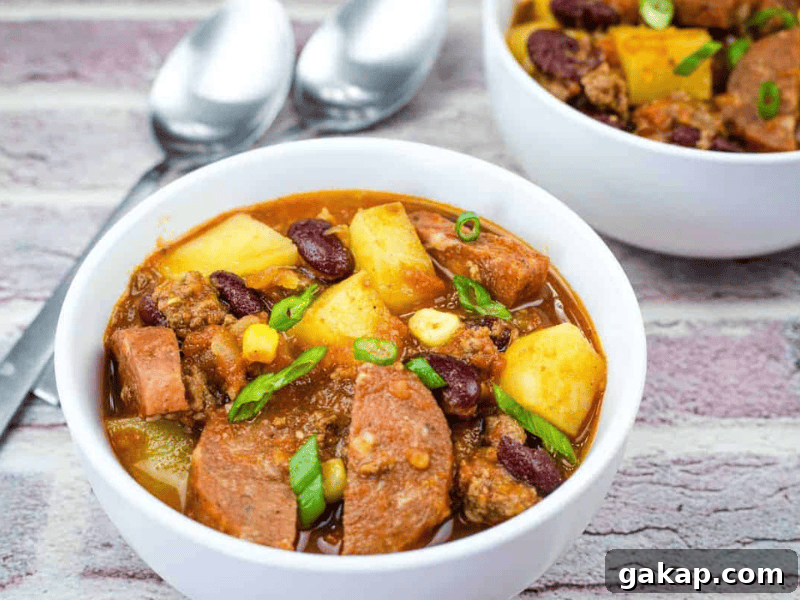 13 Classic Vintage Recipes for Cozy Fall Cooking 11 Two white bowls filled with a chunky stew of sausage, potatoes, beans, corn, and ground meat, garnished with chopped green onions. Two metal spoons are placed beside the bowls.