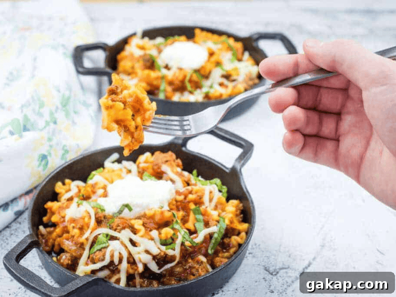 13 Classic Vintage Recipes for Cozy Fall Cooking 4 Two small cast iron pans filled with cheesy pasta and meat sauce, topped with sour cream and herbs; a hand holds a fork with a bite of pasta above one pan.