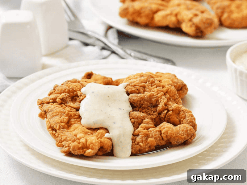 13 Classic Vintage Recipes for Cozy Fall Cooking 7 Two pieces of fried chicken steak on a white plate, topped with creamy gravy, with salt and pepper shakers and a fork in the background.