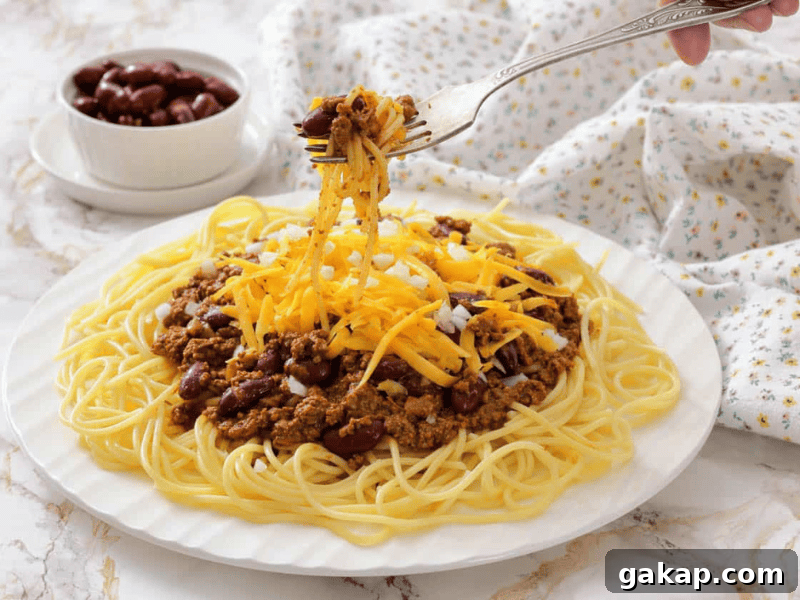 13 Classic Vintage Recipes for Cozy Fall Cooking 9 A plate of spaghetti topped with chili, shredded cheddar cheese, and chopped onions, with a fork lifting a portion; a bowl of kidney beans is in the background.
