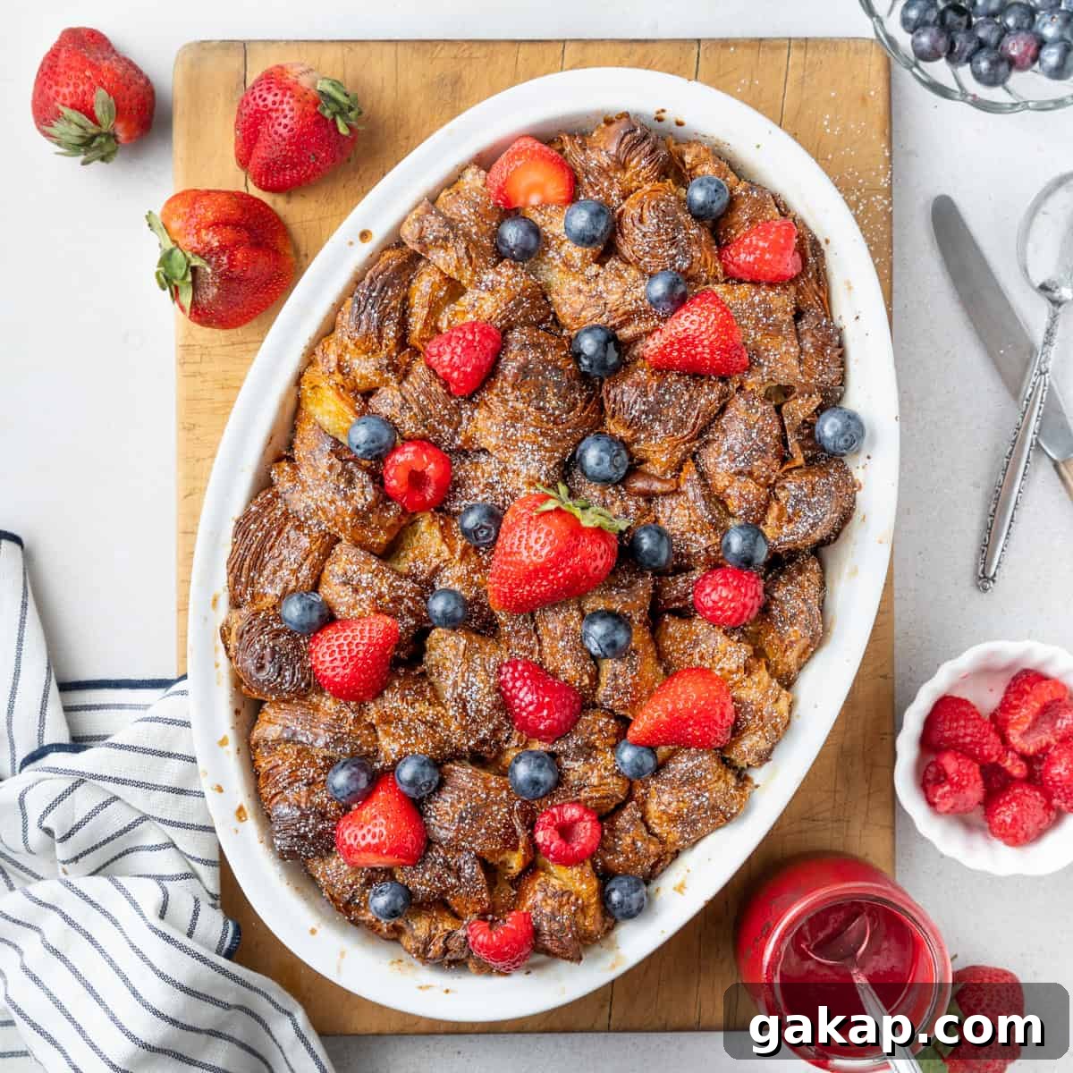 Decadent Croissant French Toast Casserole Recipe 2 French toast baked in a white oval dish seen from above, placed on a rectangular cutting board and topped with fresh berries.