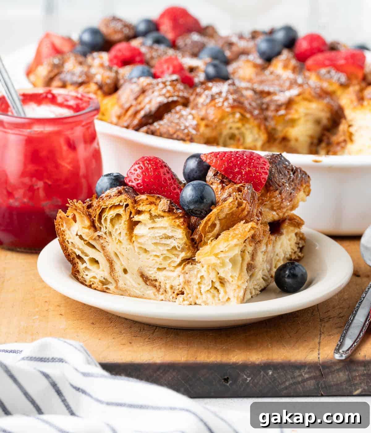 Decadent Croissant French Toast Casserole Recipe 8 Slice of french toast bake on a small white plate topped with berries, with the rest of the dish in the background.